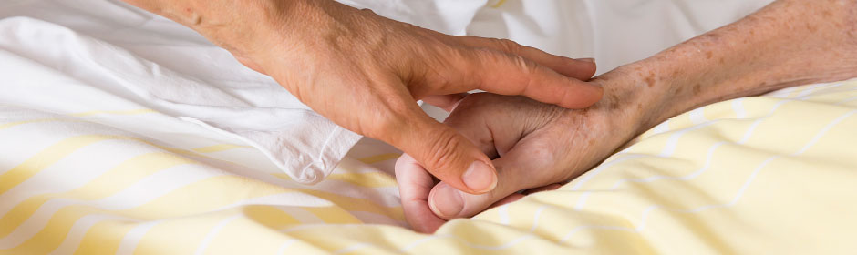 Nahaufnahme: Hand eines Menschen streicht über die einer bettlägerigen Person.