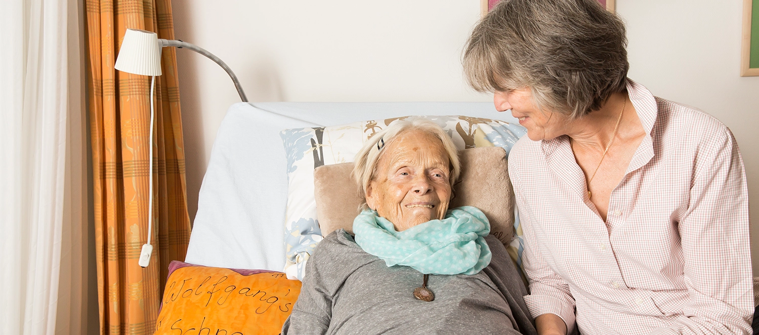 Nahaufnahme: Eine alte Frau liegt im Bett. Sie lächelt. Eine Betreuerin sitzt dicht an ihrer Seite.