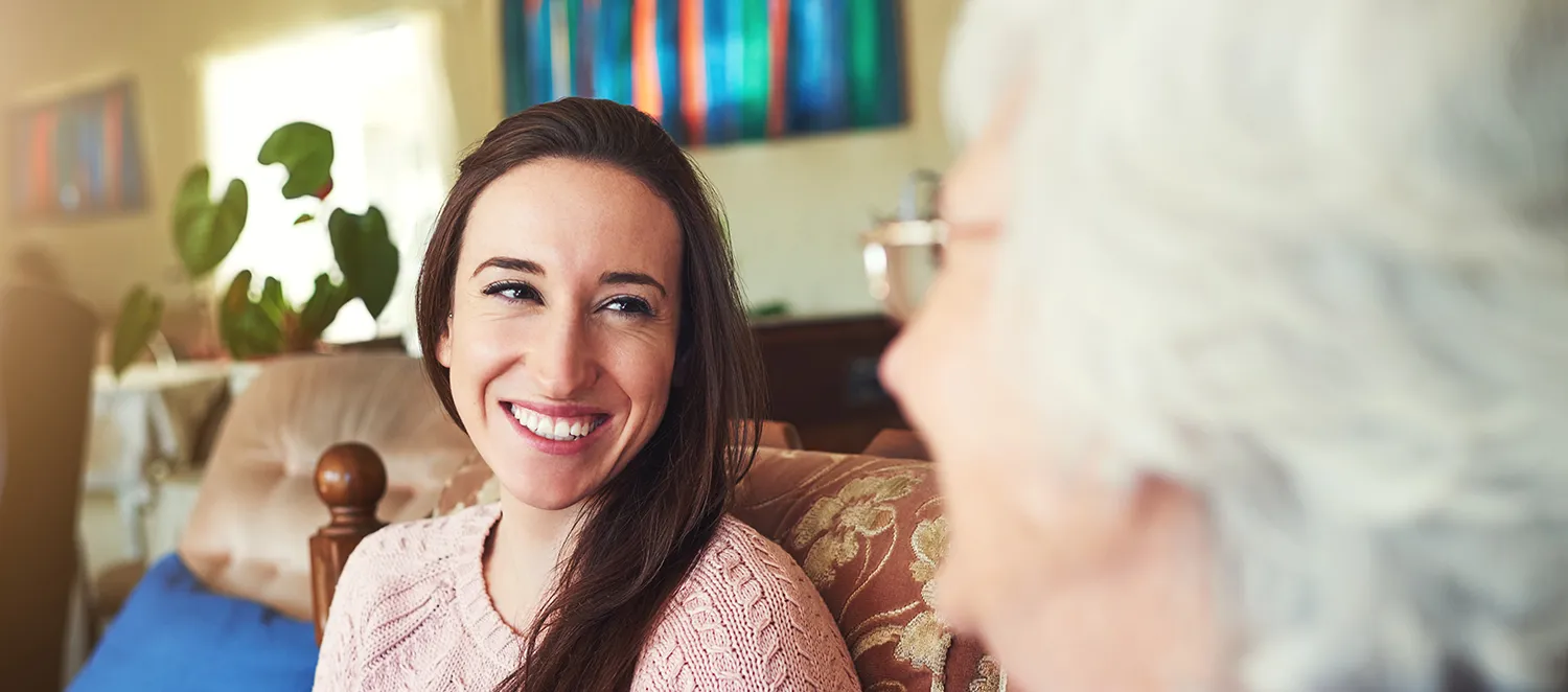 Frau sitzt mit einer Seniorin Kaffeetrinkend auf einem Sofa
