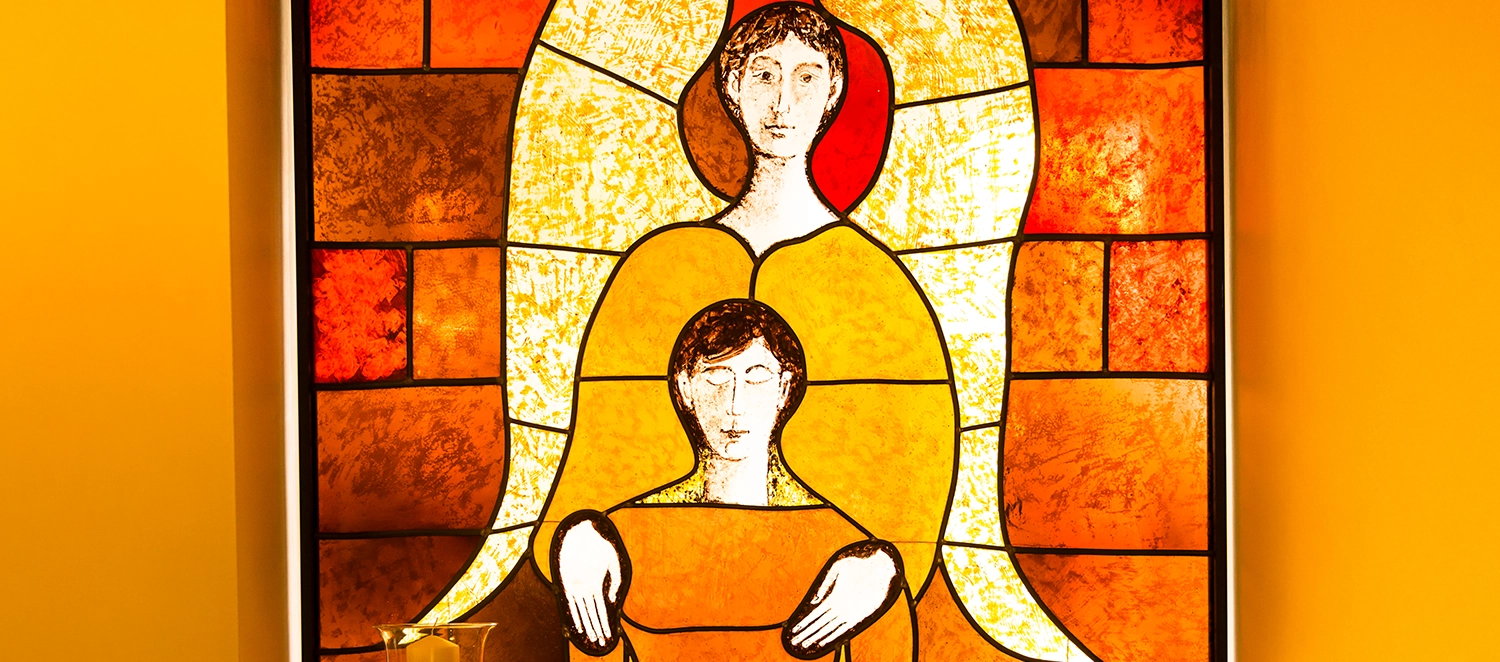 Ein Kirchenfenster mit einem Engel, welcher die Hände auf die Schultern eines Menschen legt.